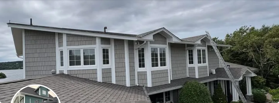A house with grey siding, white trim and a shingle roof, with a ladder set against the eave and the water behind it